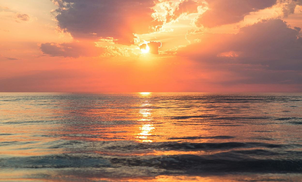 Vibrant orange and pink sunset over calm ocean waves, with the sun partially hidden behind clouds.