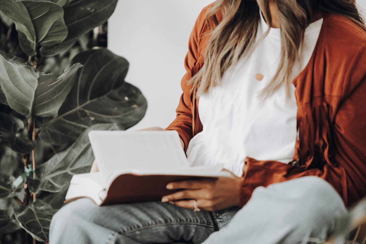 Woman sitting by a leafy plant, reading a book in her lap, wearing a white blouse and rust-colored cardigan.
