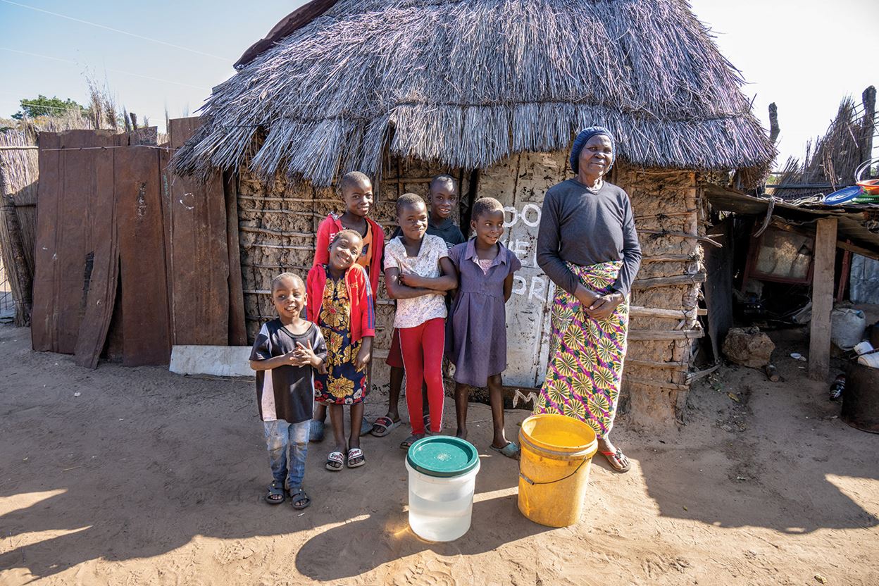 A woman and six children stand outside a thatched-roof hut, with two buckets and a large container of water placed in front of them.